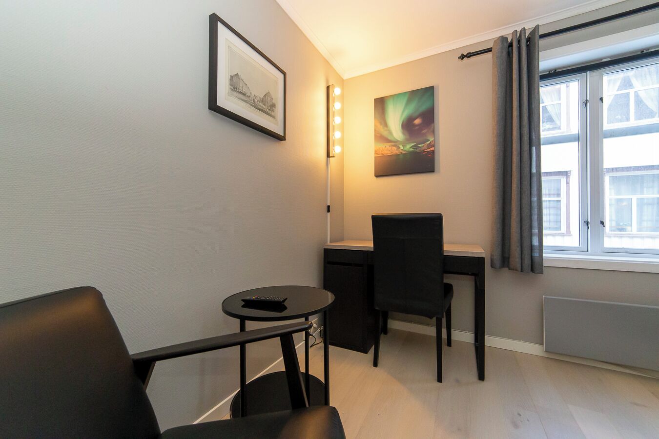 Una habitación pequeña y moderna con escritorio, silla y una mesita con un control remoto. Hay un cuadro en la pared y una ventana con cortinas.