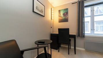 Una habitación pequeña y moderna con escritorio, silla y una mesita con un control remoto. Hay un cuadro en la pared y una ventana con cortinas.
