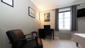 Una habitación pequeña y moderna con una silla negra, un escritorio, una cama, un radiador, una ventana con cortinas y dos cuadros en la pared.