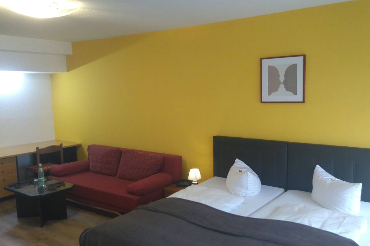 Une chambre d’hôtel avec un lit, un canapé rouge, une petite table avec une lampe et un tableau au mur.