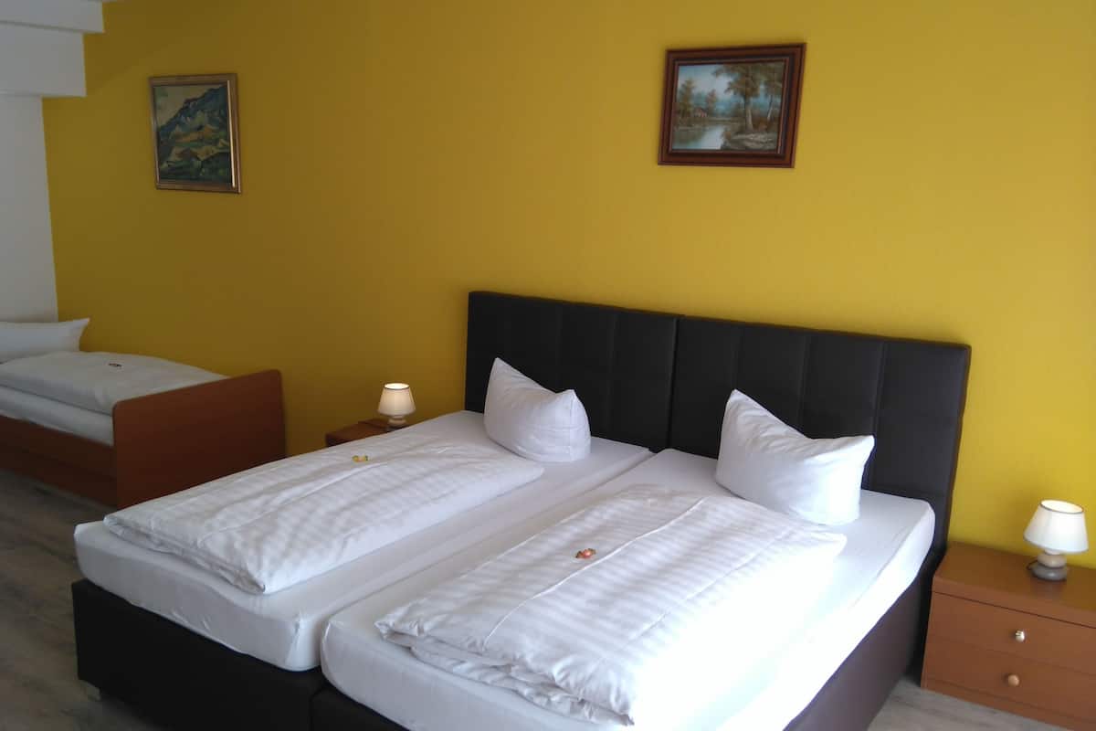 Un lit double avec du linge de lit blanc et une tête de lit sombre, un lit simple avec du linge de lit blanc, deux tables de chevet avec des lampes et deux tableaux accrochés aux murs jaunes.