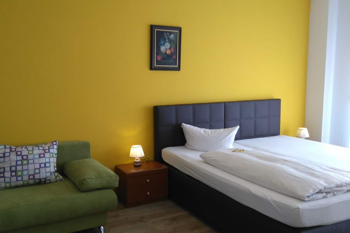 Une chambre d’hôtel avec un canapé vert, un lit avec du linge de maison blanc, une table de chevet avec une lampe et un tableau encadré au mur.
