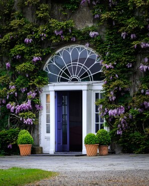 A building entrance with a white door and a fan-shaped window above it, surrounded by lush greenery and purple flowers. - Ballymaloe House Hotel (Cloyne)