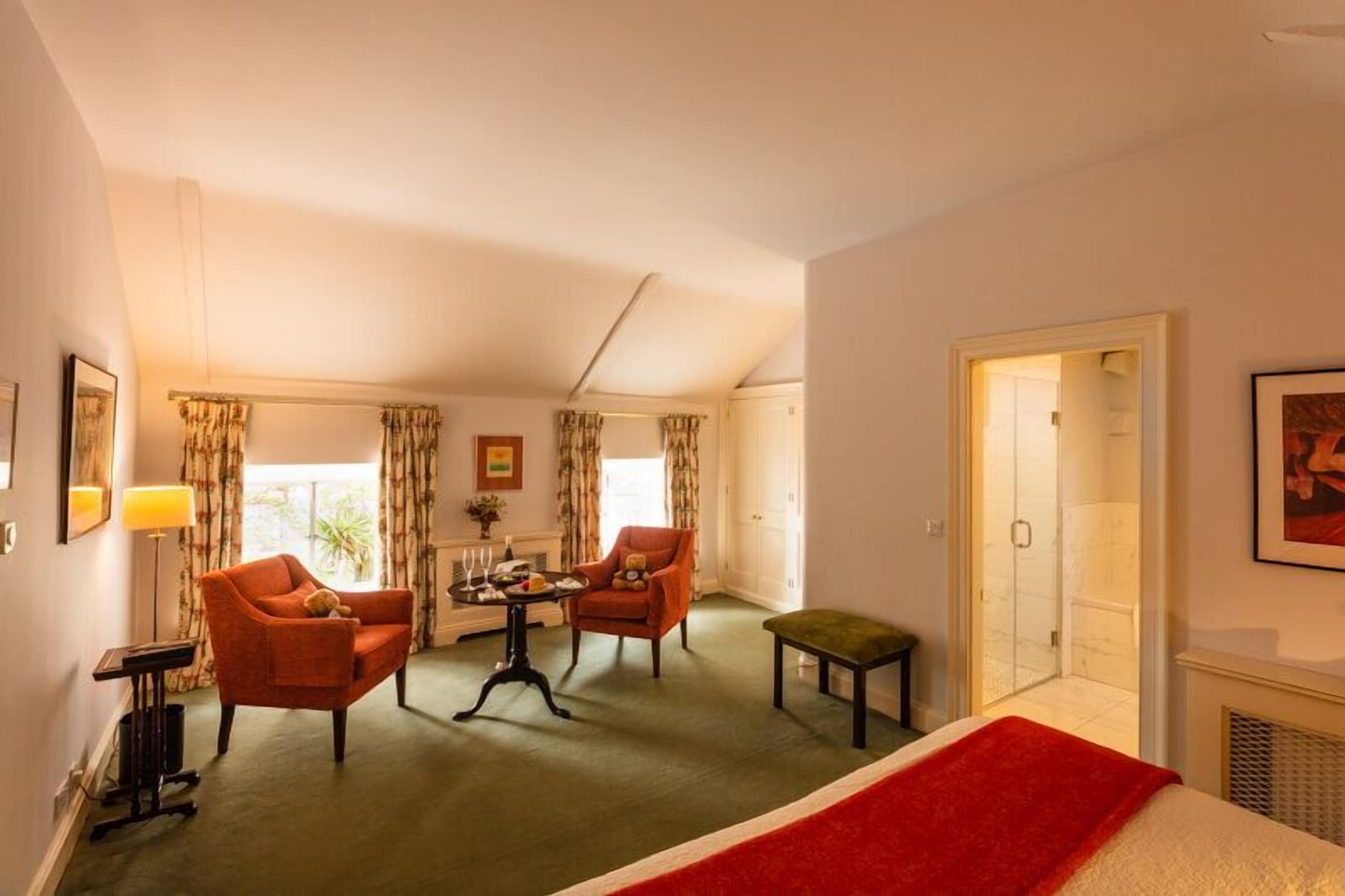 Une chambre d’hôtel comprenant un lit, deux fauteuils, une petite table, un banc, une cheminée et un espace douche.