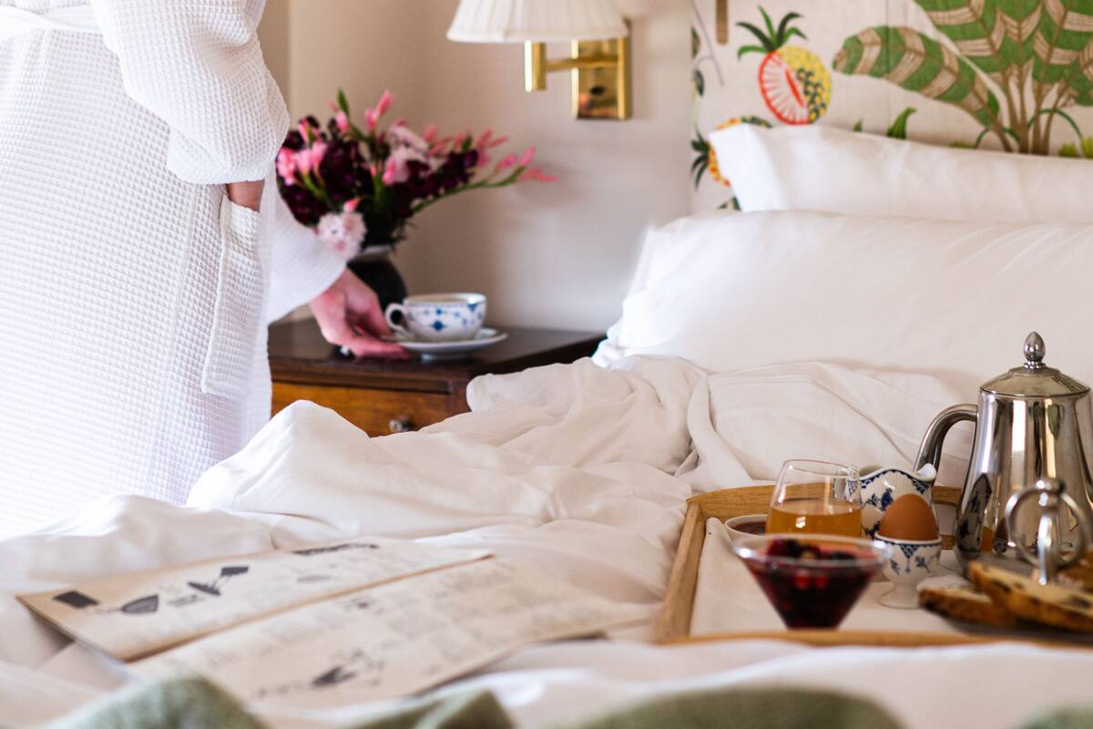 Quarto de hotel com uma cama, uma mesa de cabeceira com abajur, um tabuleiro com itens de café da manhã e uma cabeceira florida.