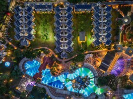 Vista aérea de um hotel com vários edifícios, área de piscina central e vegetação ao redor.