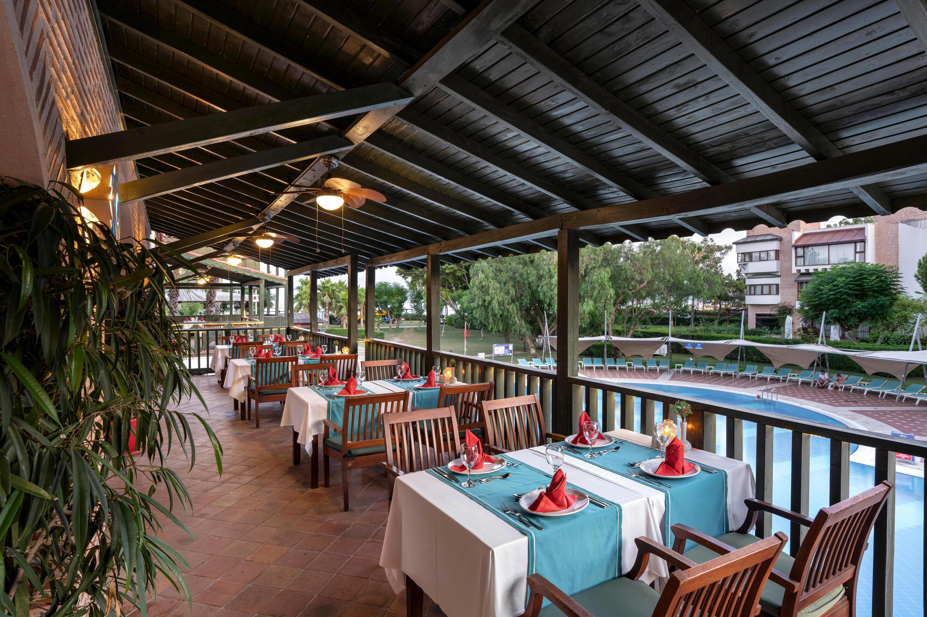 A restaurant with outdoor seating overlooking a pool, featuring wooden tables and chairs, and a covered roof structure.
