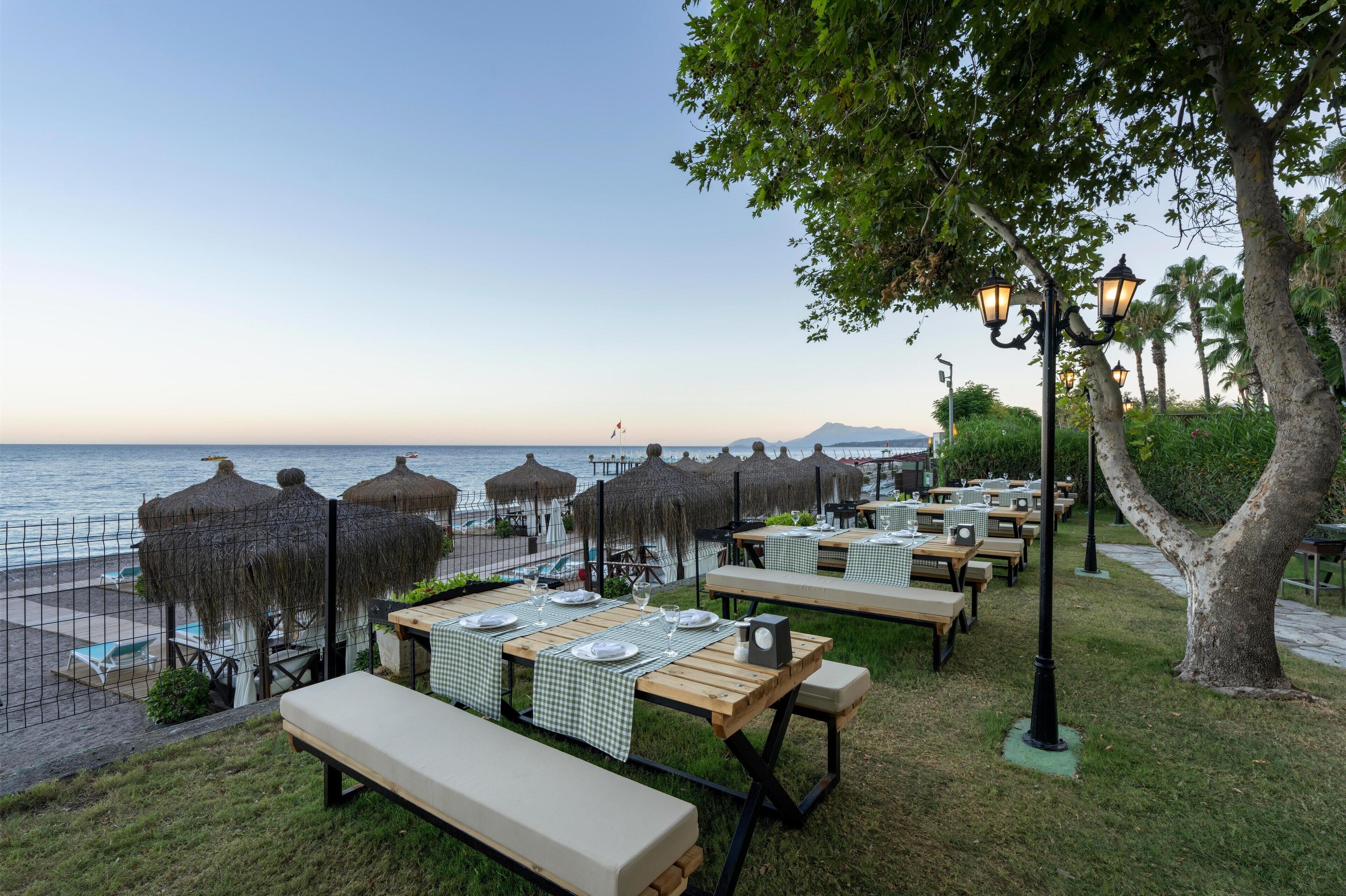 An outdoor dining area with tables set for two, overlooking a beach with umbrellas and a view of the sea.