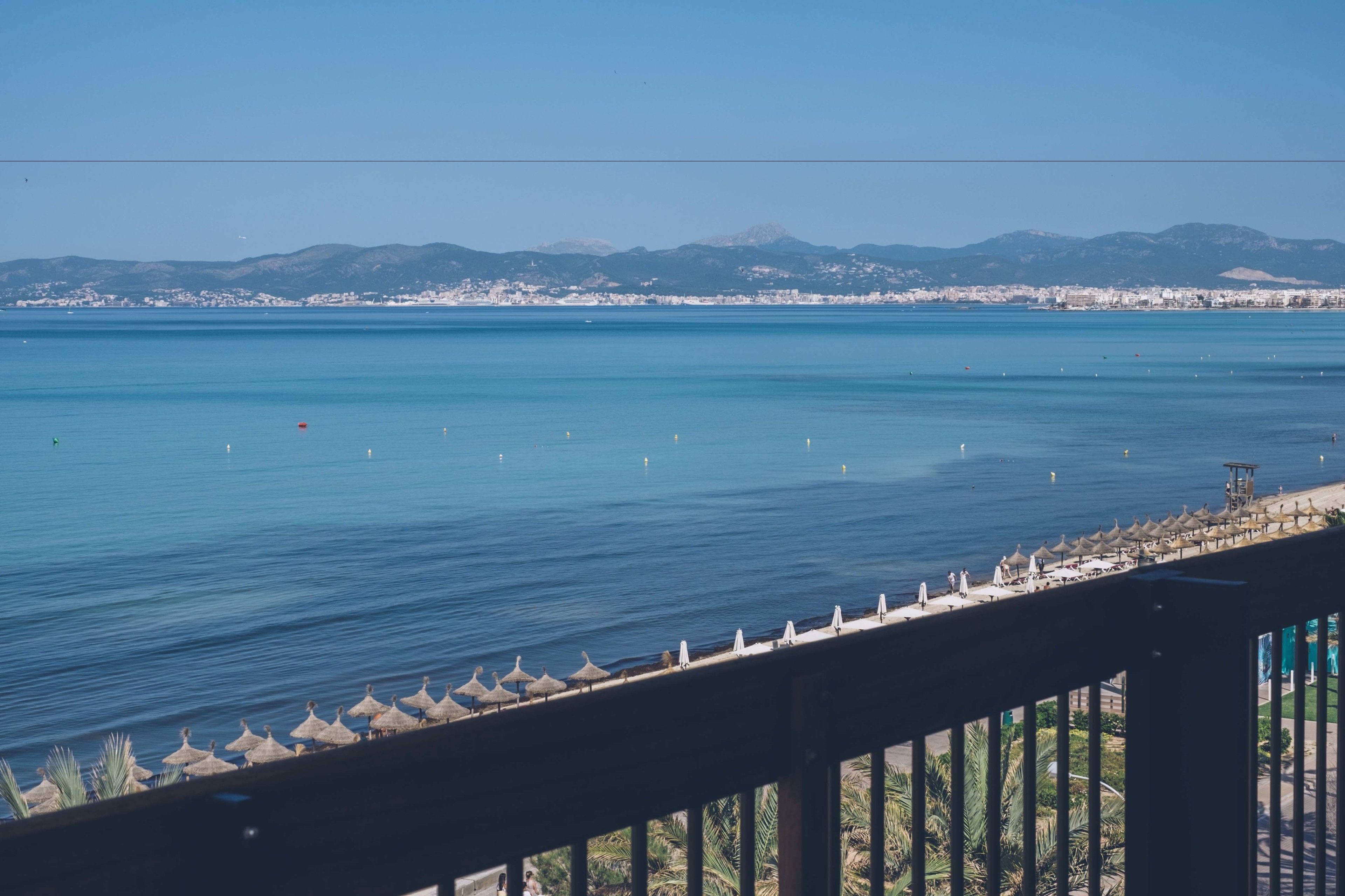 Vista costera con mar azul cristalino, un paseo marítimo con sombrillas y montañas a lo lejos.