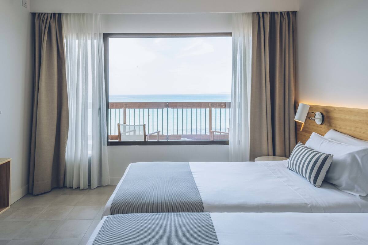 Ein Hotelzimmer mit zwei Betten, einem großen Fenster mit Meerblick und einem Balkon mit Geländer.