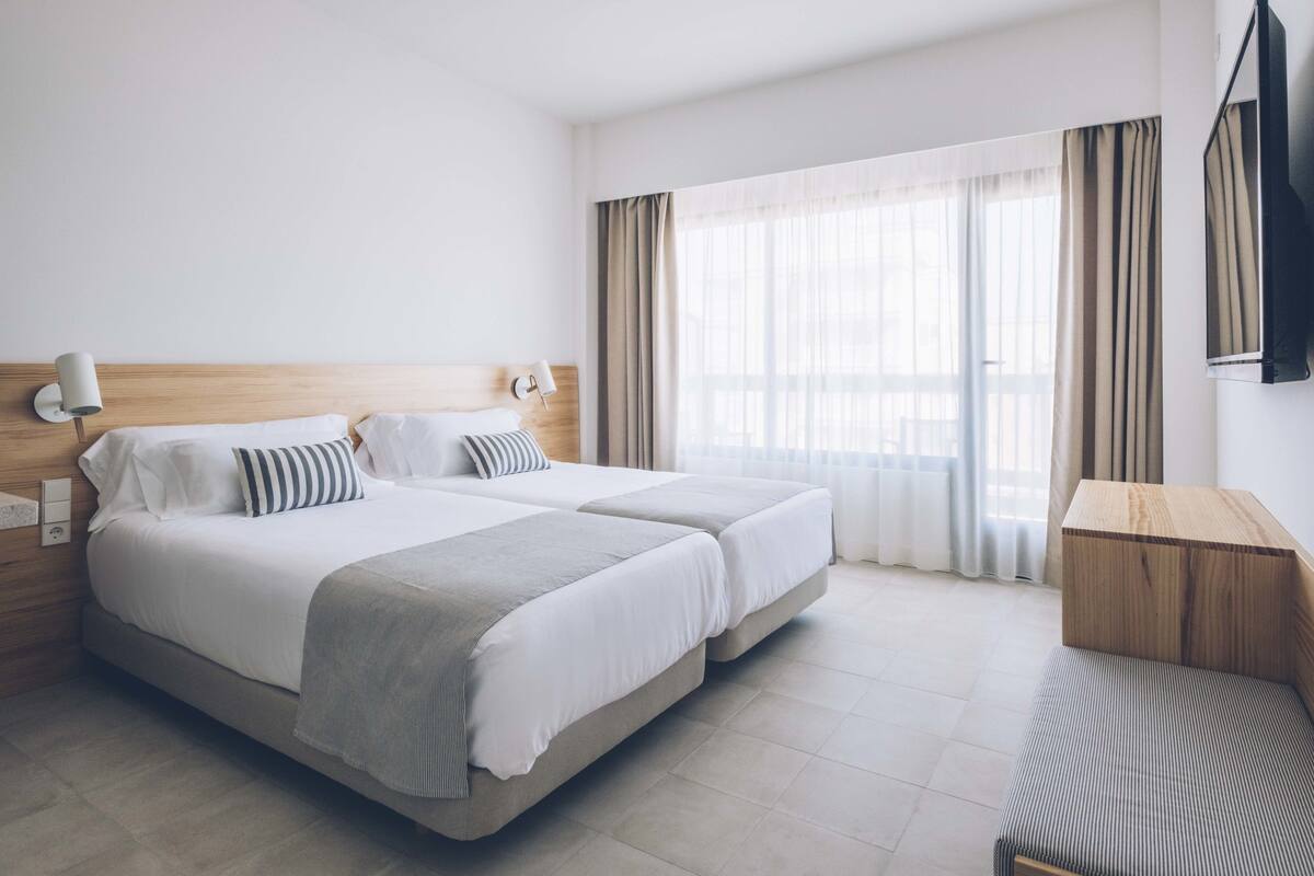 Ein Hotelzimmer mit zwei Betten, einem hölzernen Kopfteil, einem Nachttisch mit Lampe, einem großen Fenster mit Vorhängen und einer Holzbank.