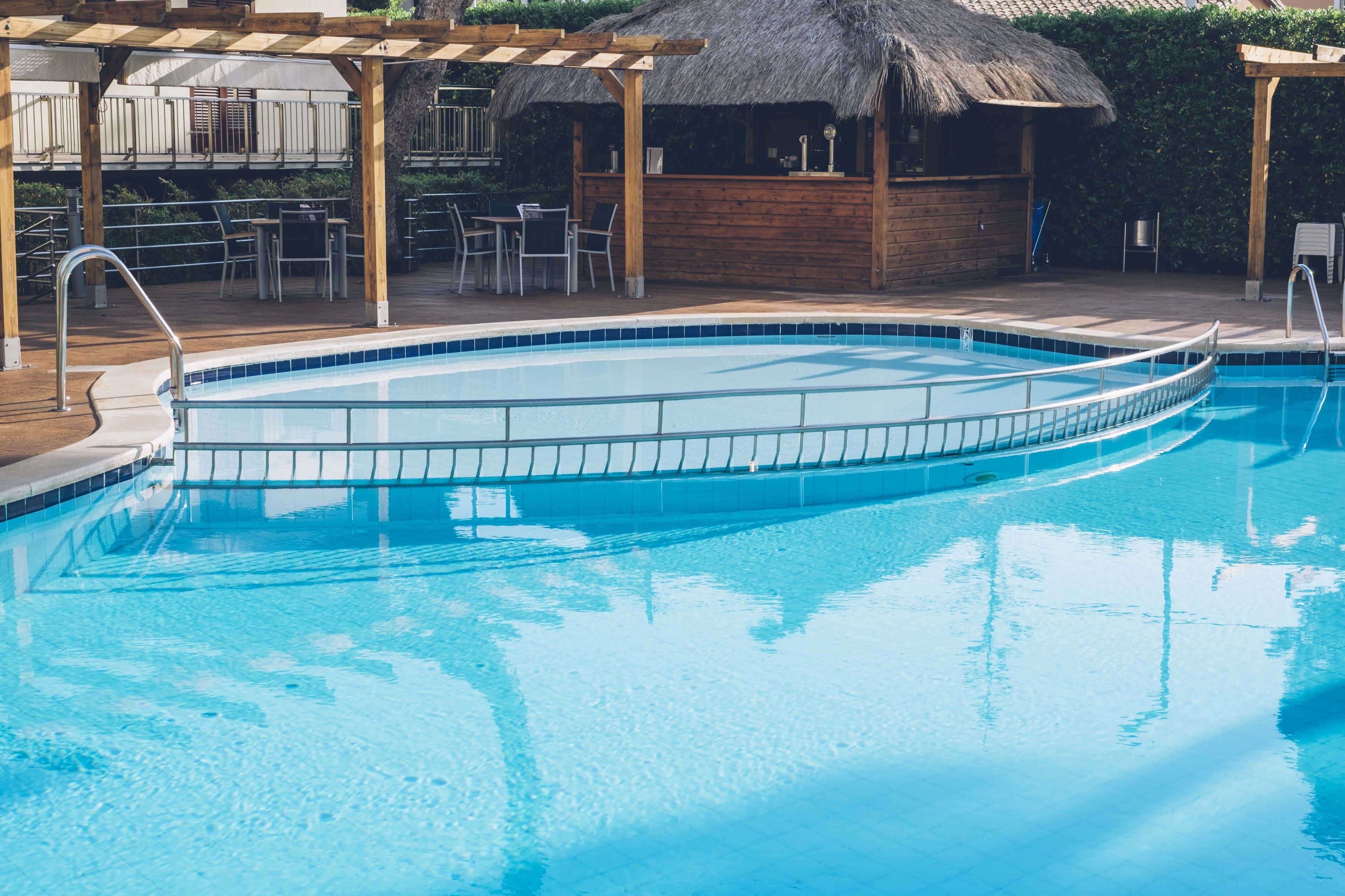 Una piscina con borde de azulejos, cubierta de madera y una estructura de techo de paja al fondo.