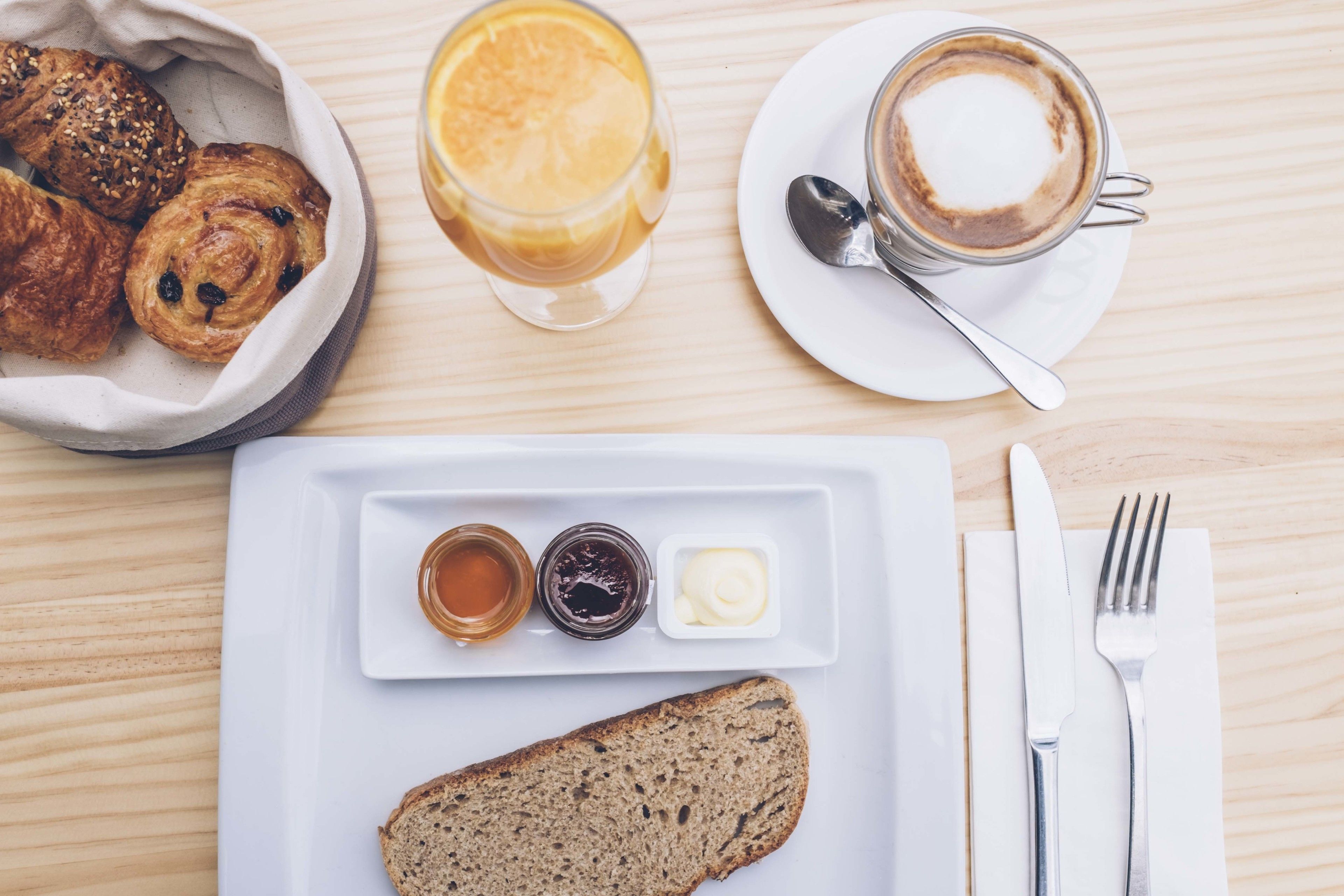 Un desayuno con cruasanes, un vaso de jugo de naranja, una taza de café, una rebanada de pan y condimentos sobre un plato blanco.