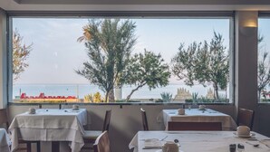Un restaurante con mesas dispuestas para comer, vista al mar y árboles fuera de la ventana.