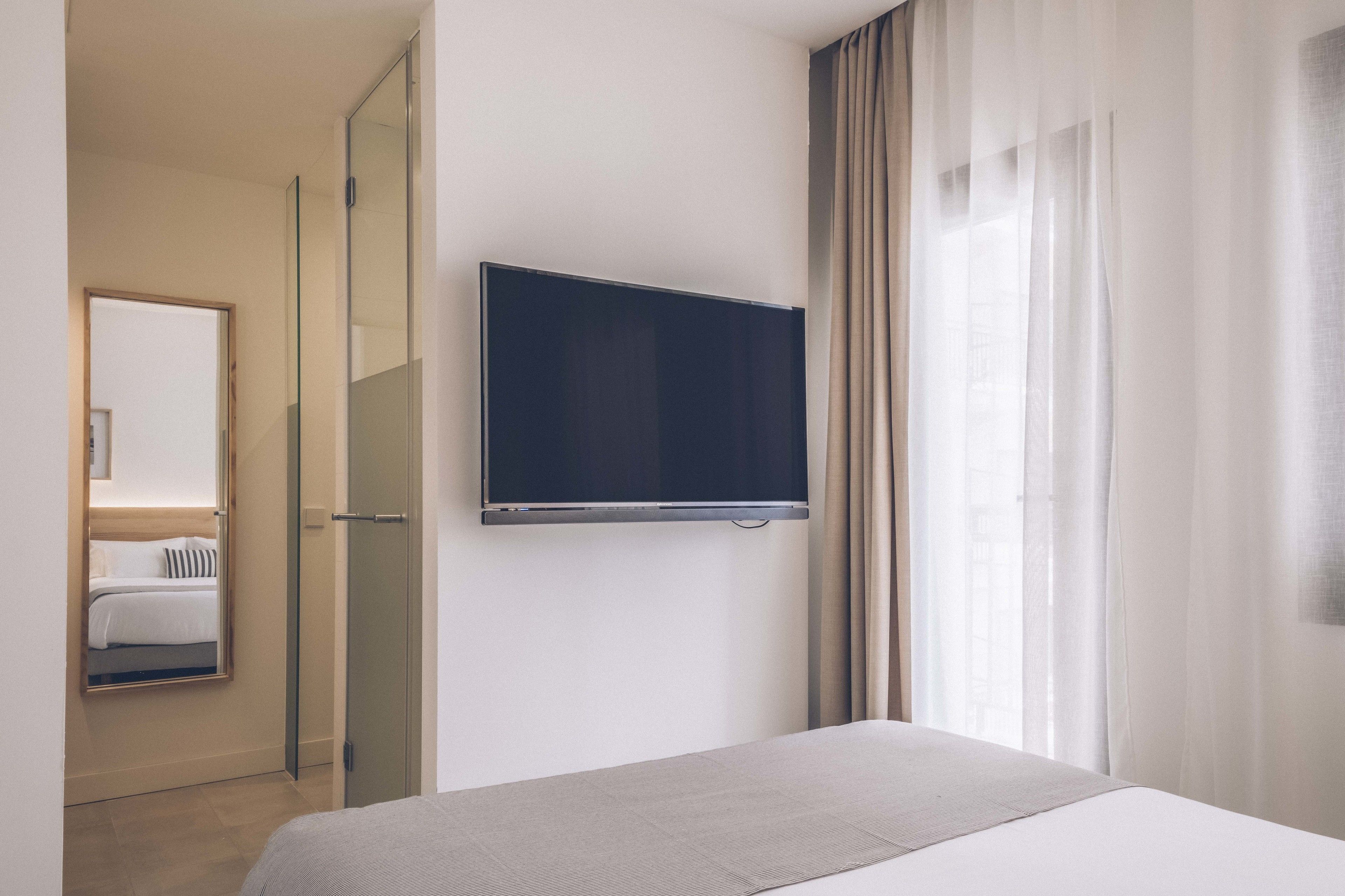 Ein Hotelzimmer mit Bett, Wandfernseher, Glastür zum Badezimmer und einem blickdichten Fenster.
