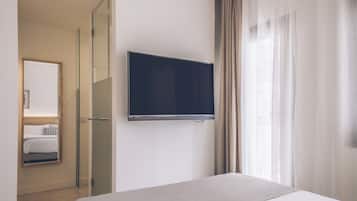 Ein Hotelzimmer mit Bett, Wandfernseher, Glastür zum Badezimmer und einem blickdichten Fenster.