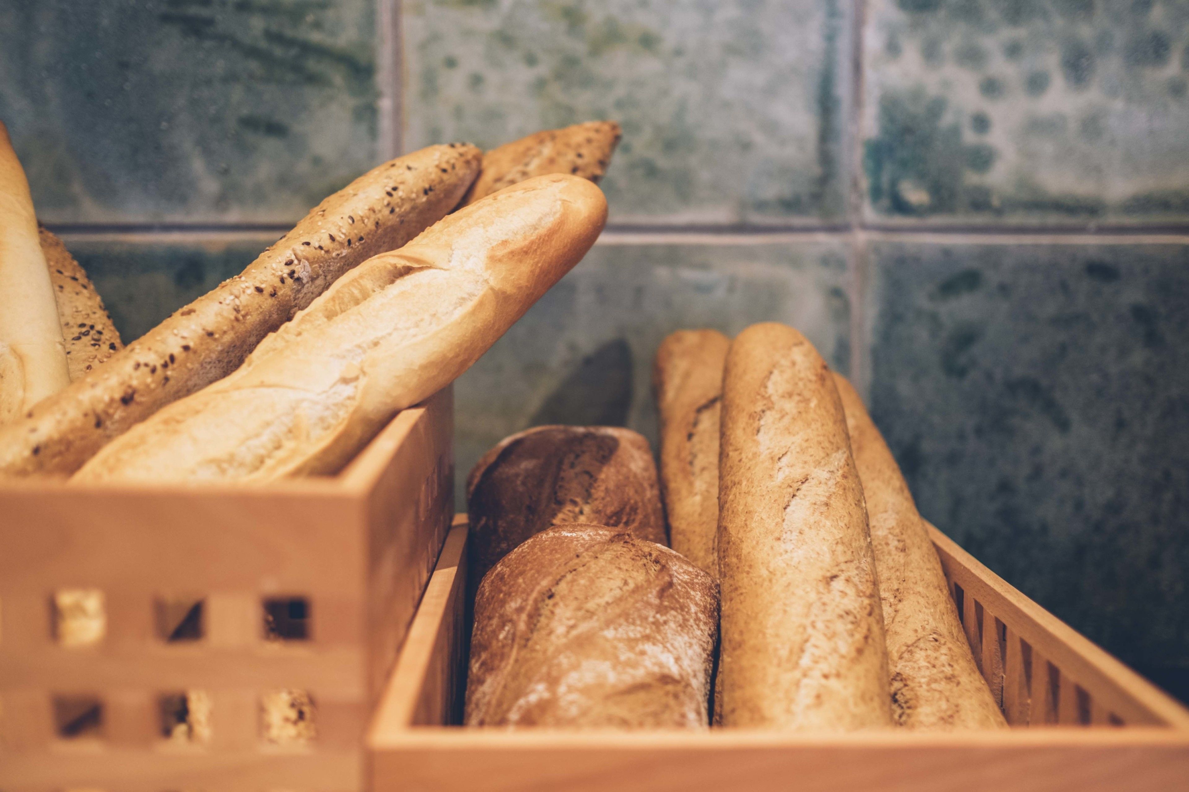 A wooden crate filled with various types of bread, including baguettes and rolls, against a tiled wall background.