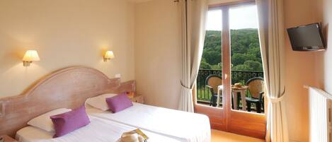 A double bed with white linens and purple pillows, a ceiling fan, a TV on the wall, and a balcony with outdoor furniture.