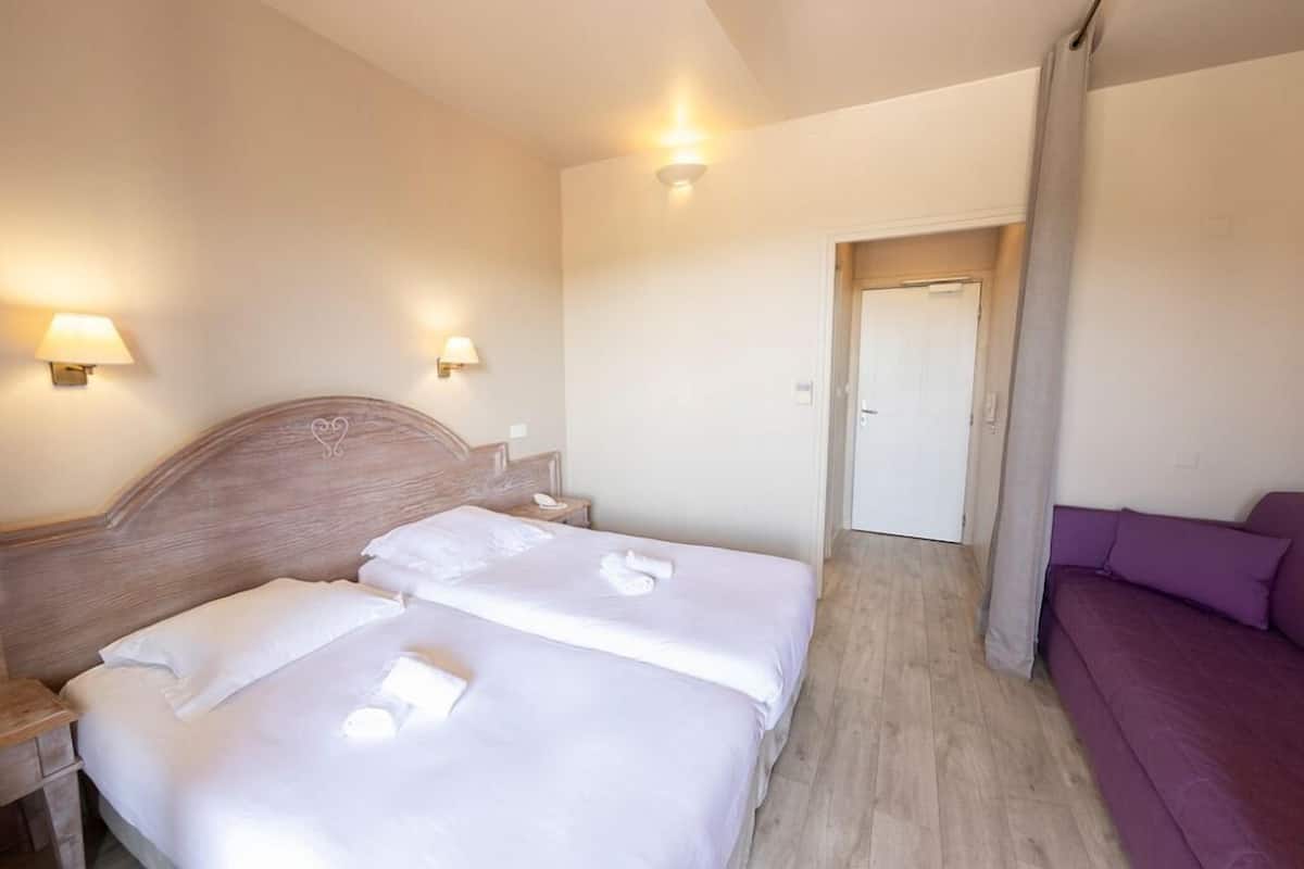 Habitación de hotel con dos camas, cabecero de madera, lámparas de noche, sofá morado y una puerta que da a otra habitación.