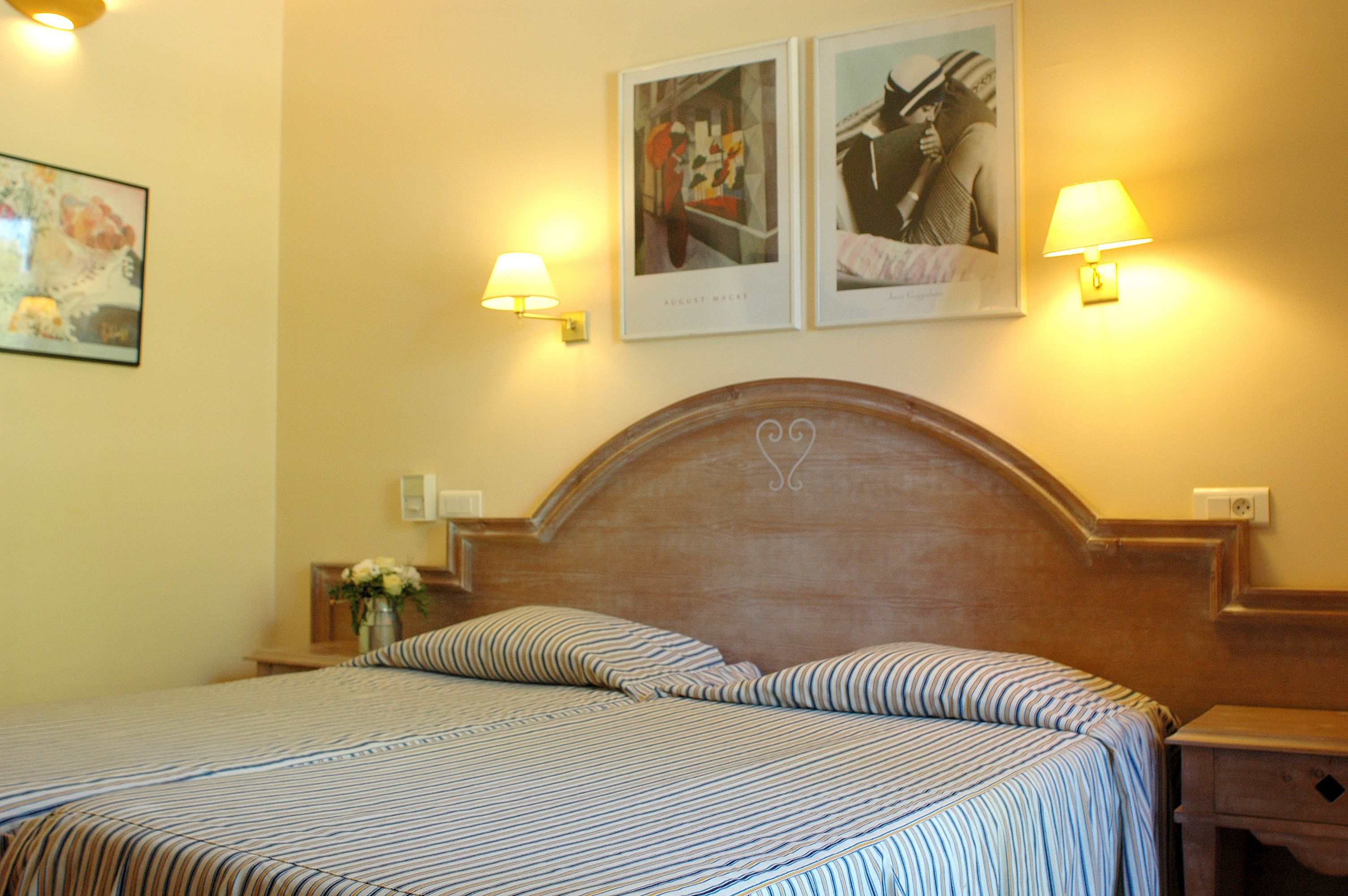 A hotel room with a bed, two wall-mounted lamps, and two framed pictures on the wall.