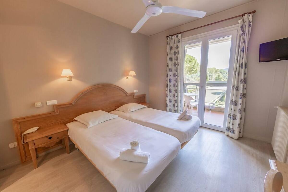 Habitación de hotel con dos camas, una mesita de noche de madera, un escritorio, un ventilador de techo y un balcón con cortinas.