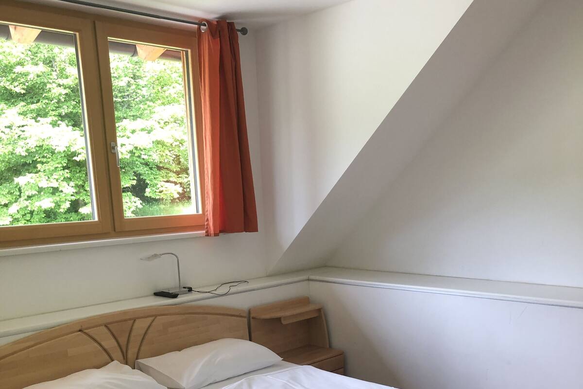 Una camera da letto con un letto di legno, un comodino, una finestra con tende e vista sul verde.