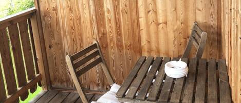 A wooden deck with a table and two chairs, a white cushion on one chair, and a view of a green landscape.
