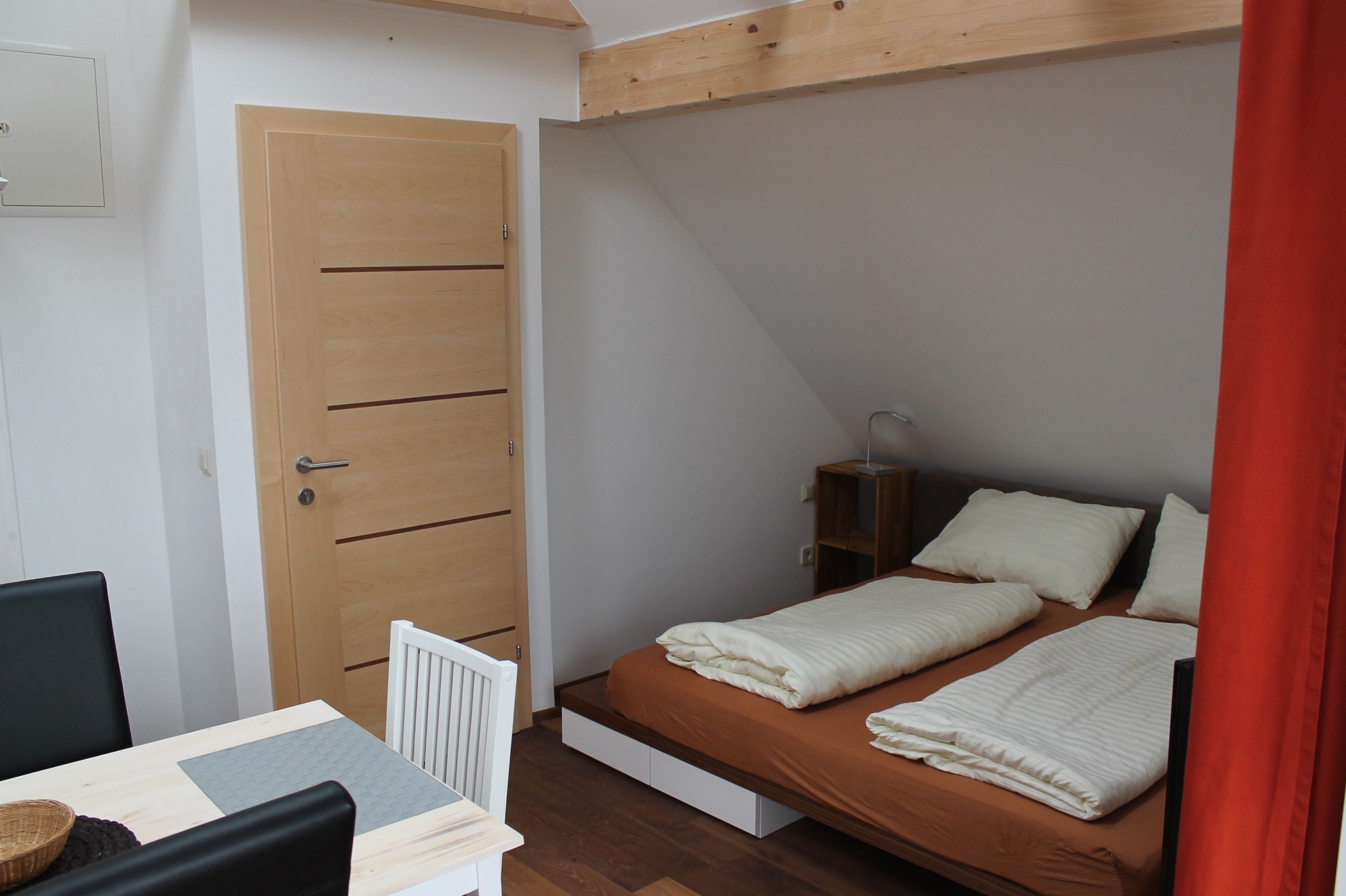 Une petite chambre bien éclairée, avec un lit simple, une porte en bois, une petite table avec une chaise et un radiateur.
