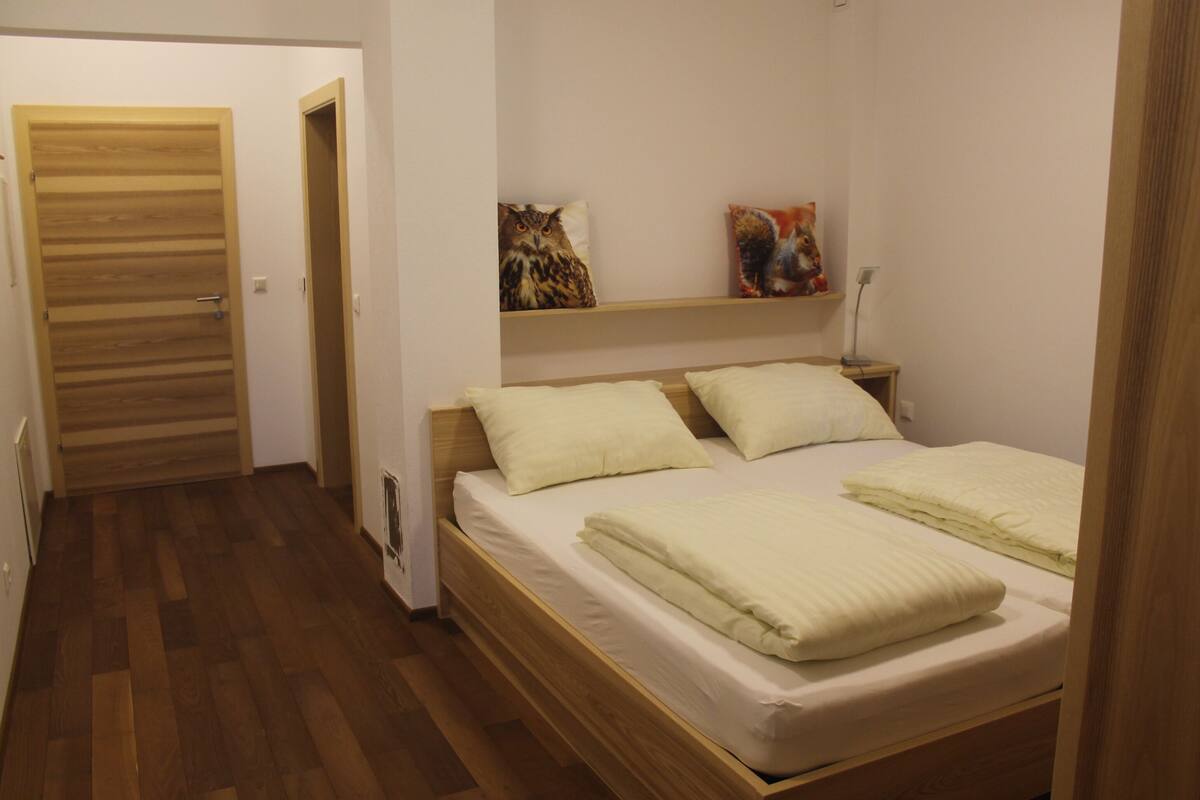 Una camera da letto con un letto di legno, biancheria bianca, una porta in legno e uno scaffale con due cuscini decorativi.