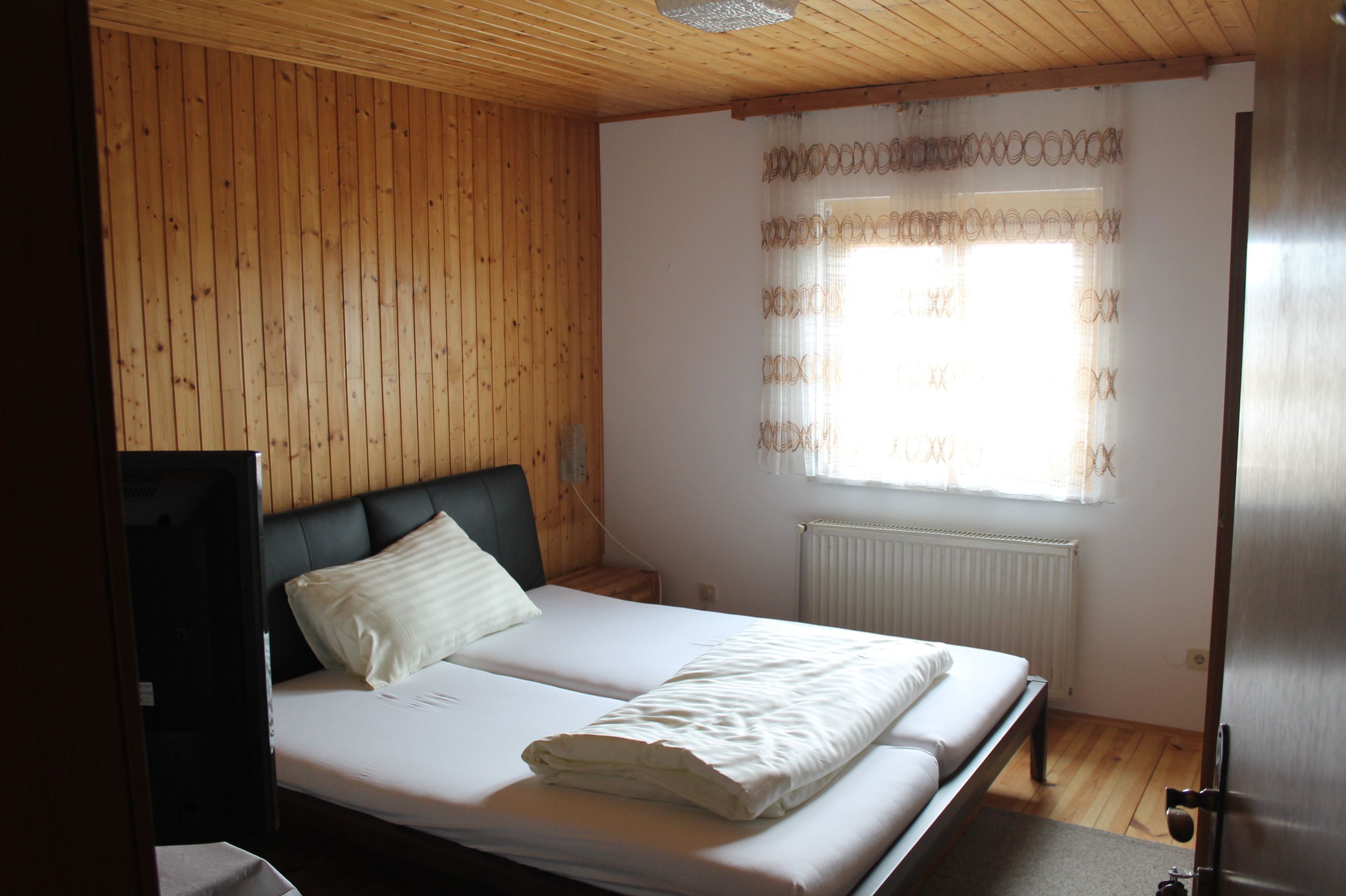 Ein Zimmer mit einer Holzvertäfelung, einem Bett mit weißer Bettwäsche, einem Nachttisch und einem Fenster mit gemusterten Vorhängen.