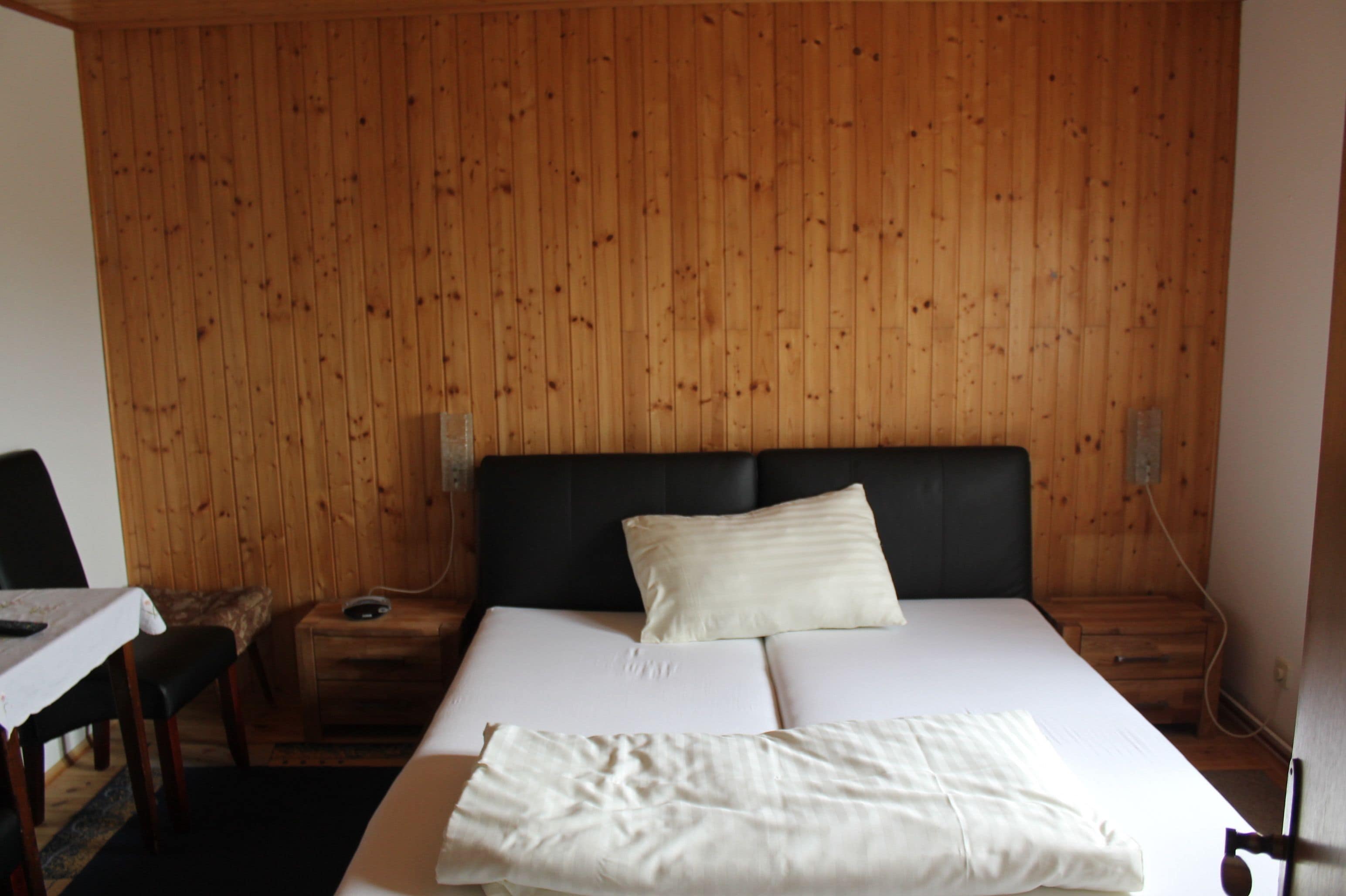 A room with a wooden paneled wall, a bed with white bedding, a bedside table, and a chair.
