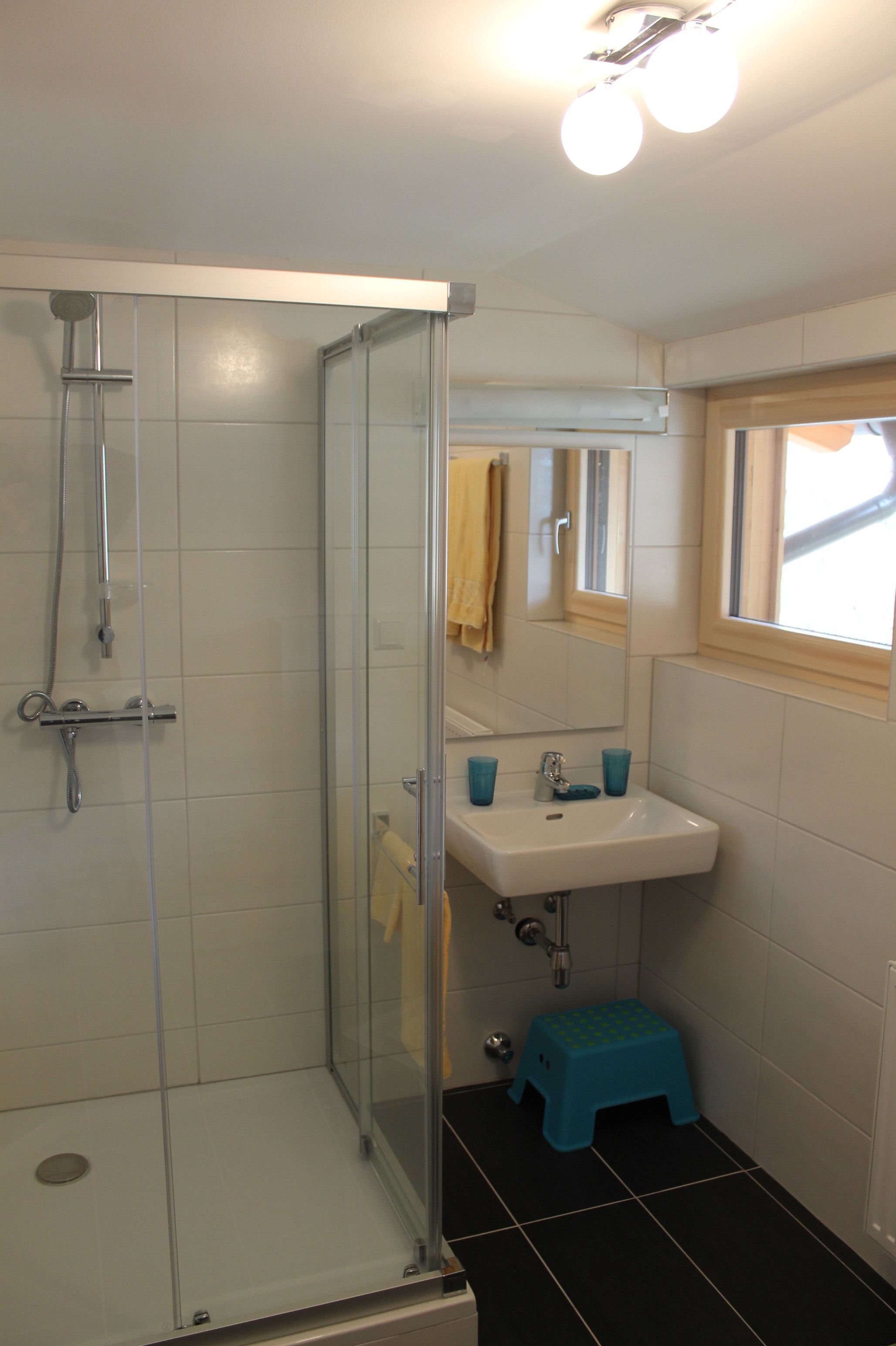 Een badkamer met een douchecabine van glas, een wastafel met twee blauwe bekers en een klein blauw krukje.