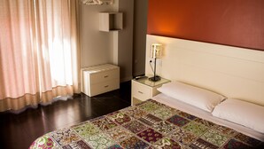 Une chambre d’hôtel avec un lit, une table de chevet avec une lampe, une commode et une fenêtre avec des rideaux.