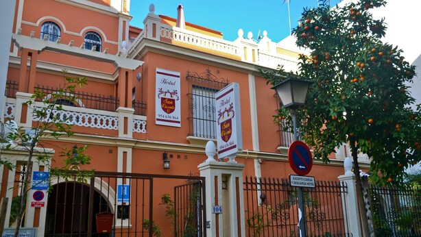 La façade d’un hôtel aux couleurs rouge et blanc, avec une entrée fermée par un portail et un panneau indiquant qu’il est interdit de se garer.