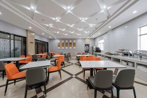 Ένας χώρος εστίασης με τραπέζια και καρέκλες, ένας μπουφές και μια κουζίνα στο βάθος.