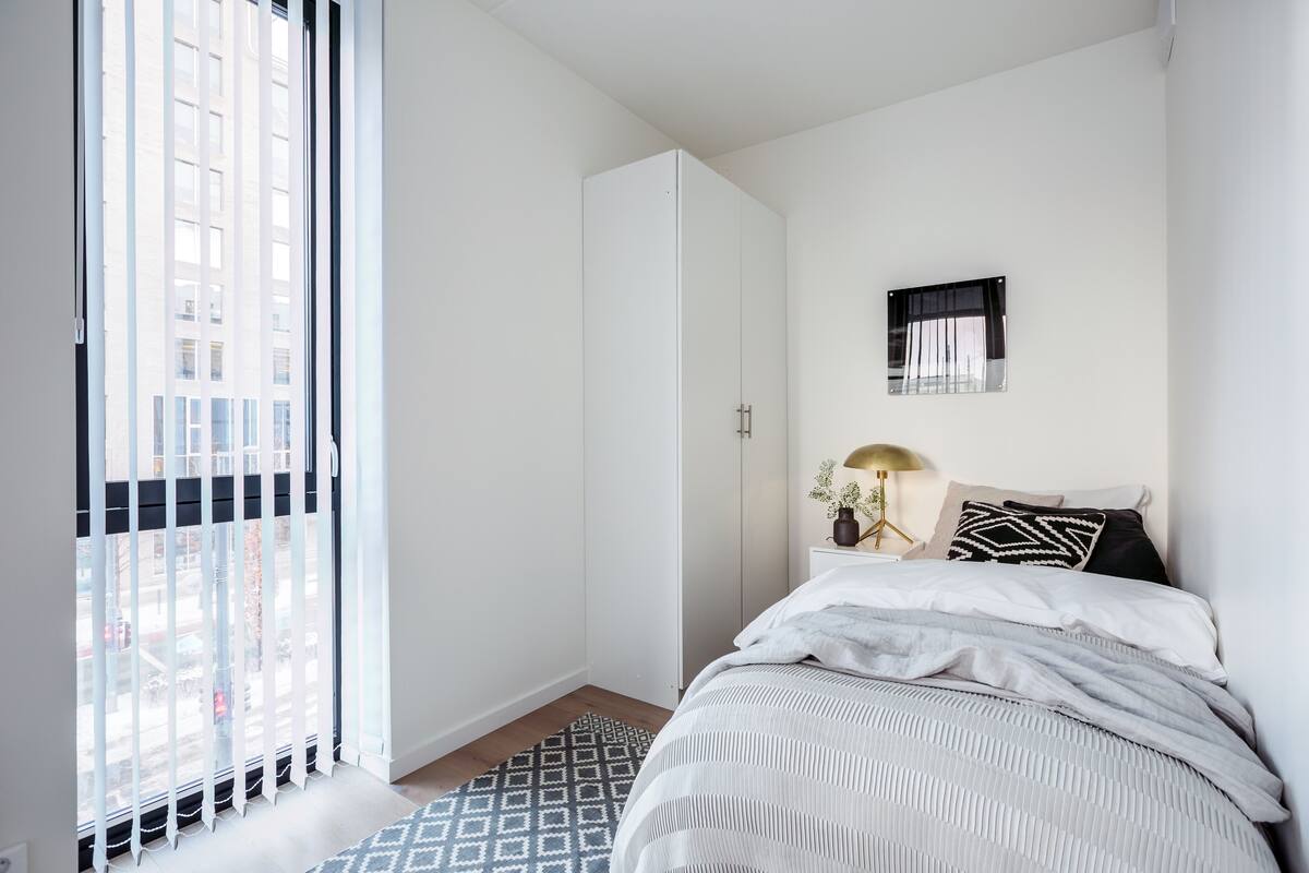 A bedroom with a bed, a wardrobe, a window with blinds, and a small bedside table with a lamp and a plant.