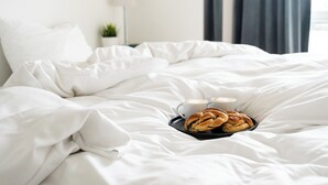 Un lit bien fait, avec du linge de lit blanc, un plateau avec des pâtisseries et deux tasses de café, et une lampe en arrière-plan.
