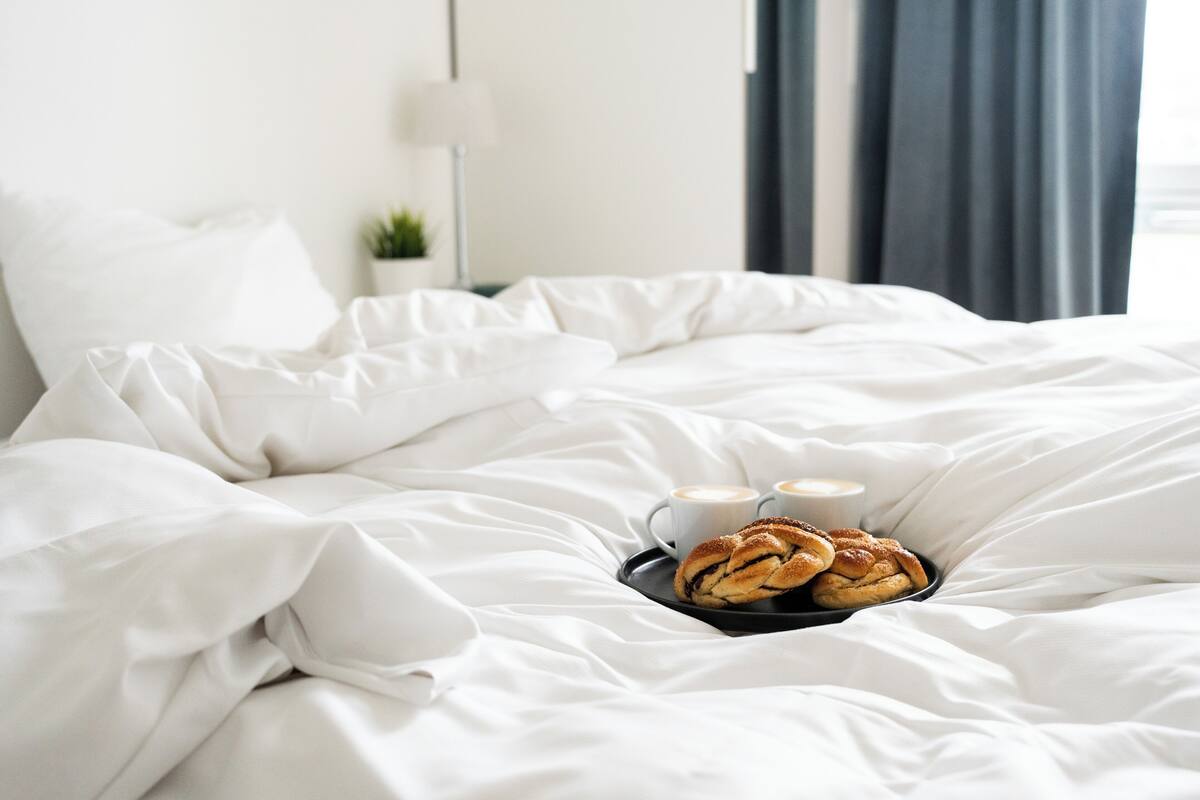 A neatly made bed with white linens, a tray with pastries and two cups of coffee, and a lamp in the background.