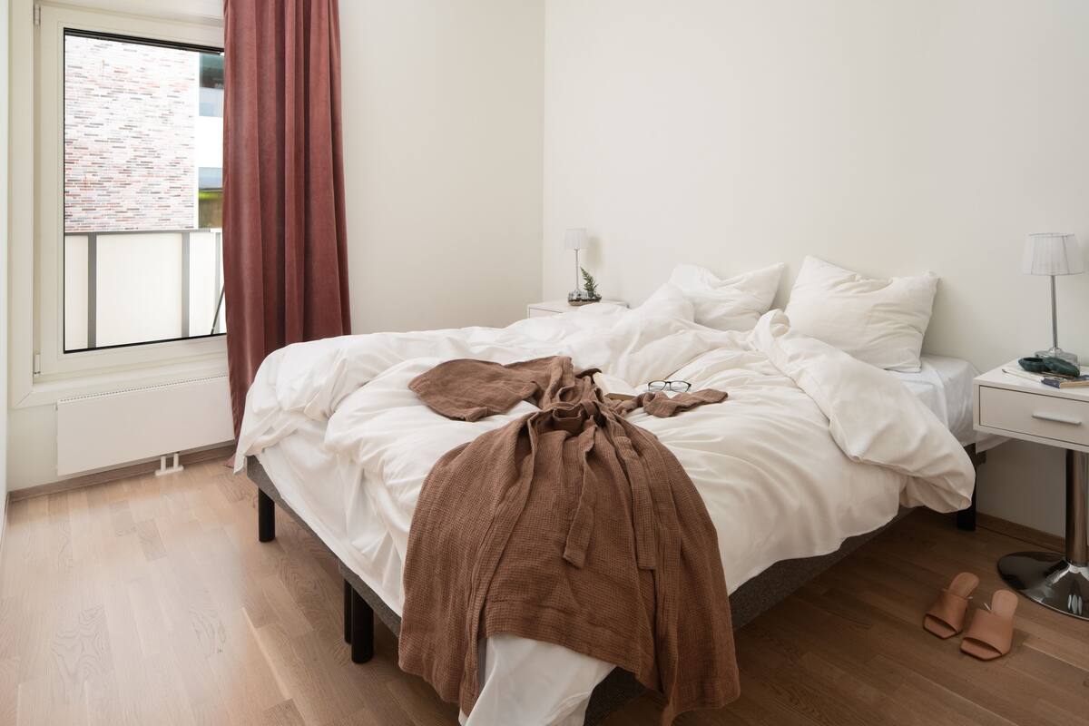 A neatly made bed with a brown blanket, a bedside table with a lamp, and a pair of sandals on the floor.
