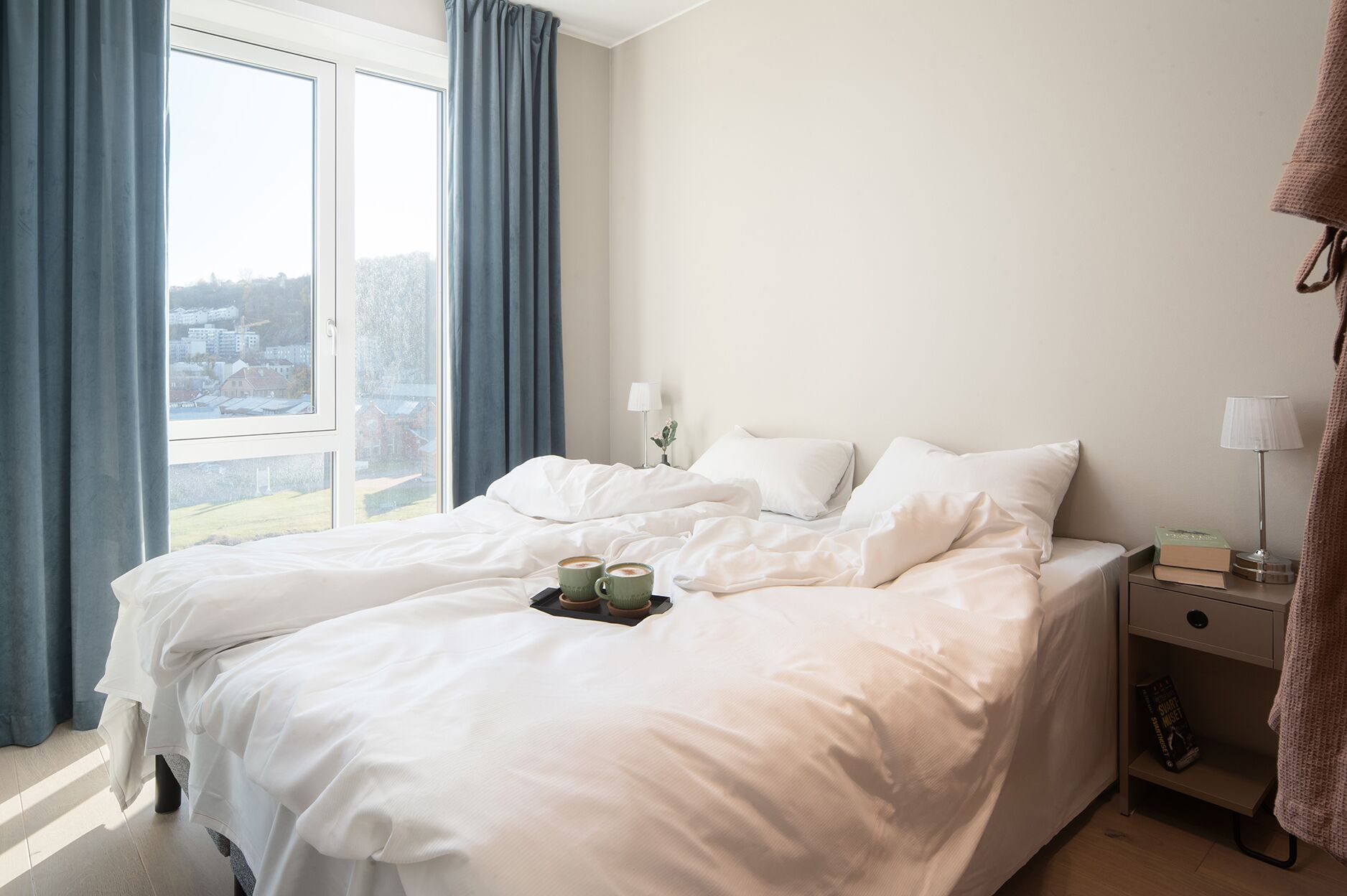 A hotel room with a large bed, a bedside table with a lamp, and a view of a cityscape through the window.