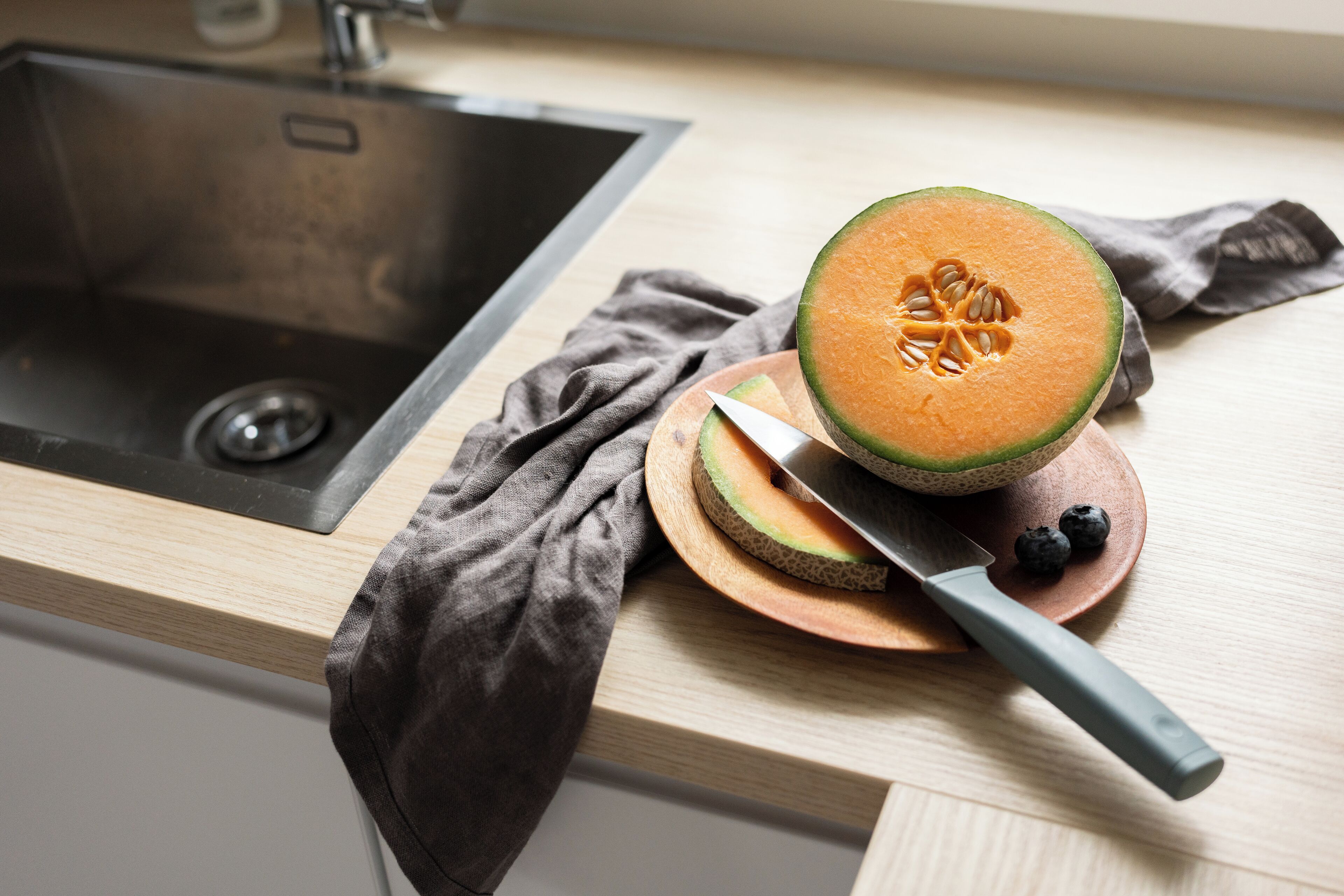 Una encimera de cocina con un lavabo, un melón partido, un cuchillo y un paño.