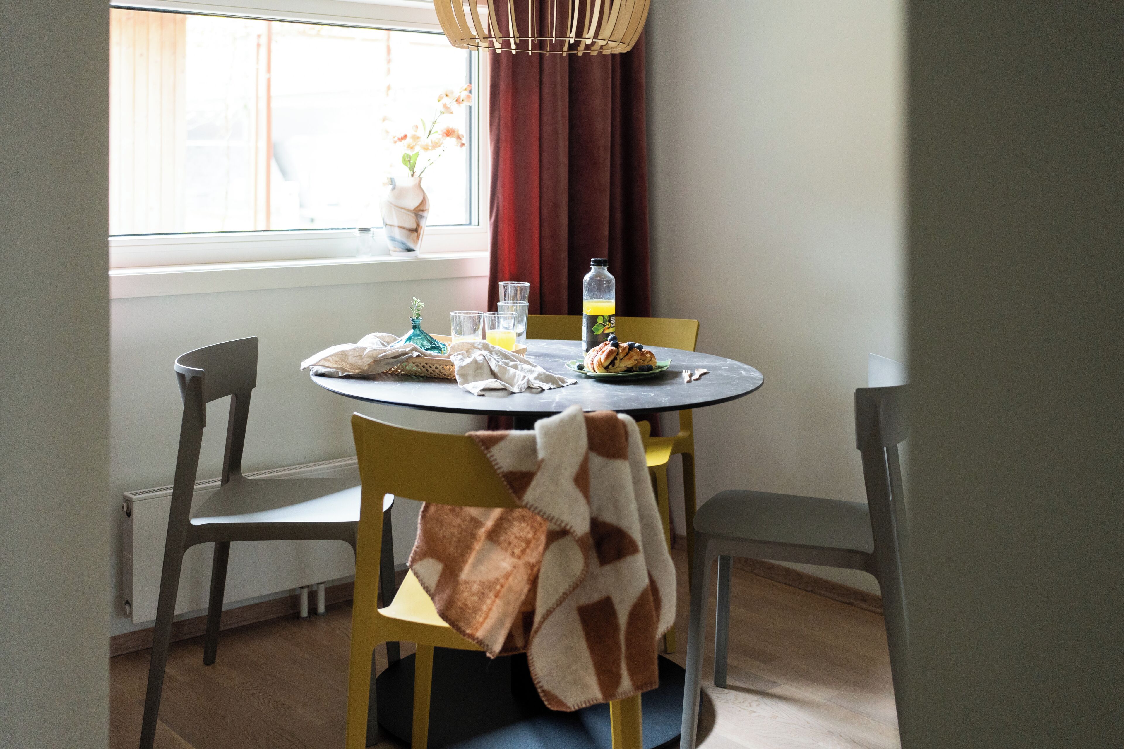 Een eetgedeelte met een ronde tafel, stoelen, een vaas met bloemen en een fles sinaasappelsap.