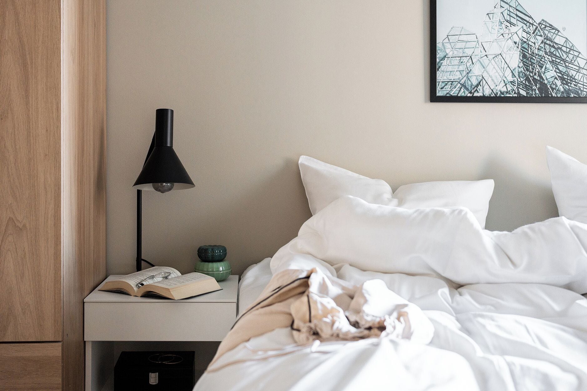 A neatly made bed with white bedding, a bedside table with a lamp, a book, and a small green bowl.