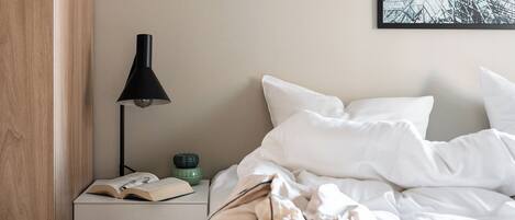 A neatly made bed with white bedding, a bedside table with a lamp, a book, and a small green bowl.