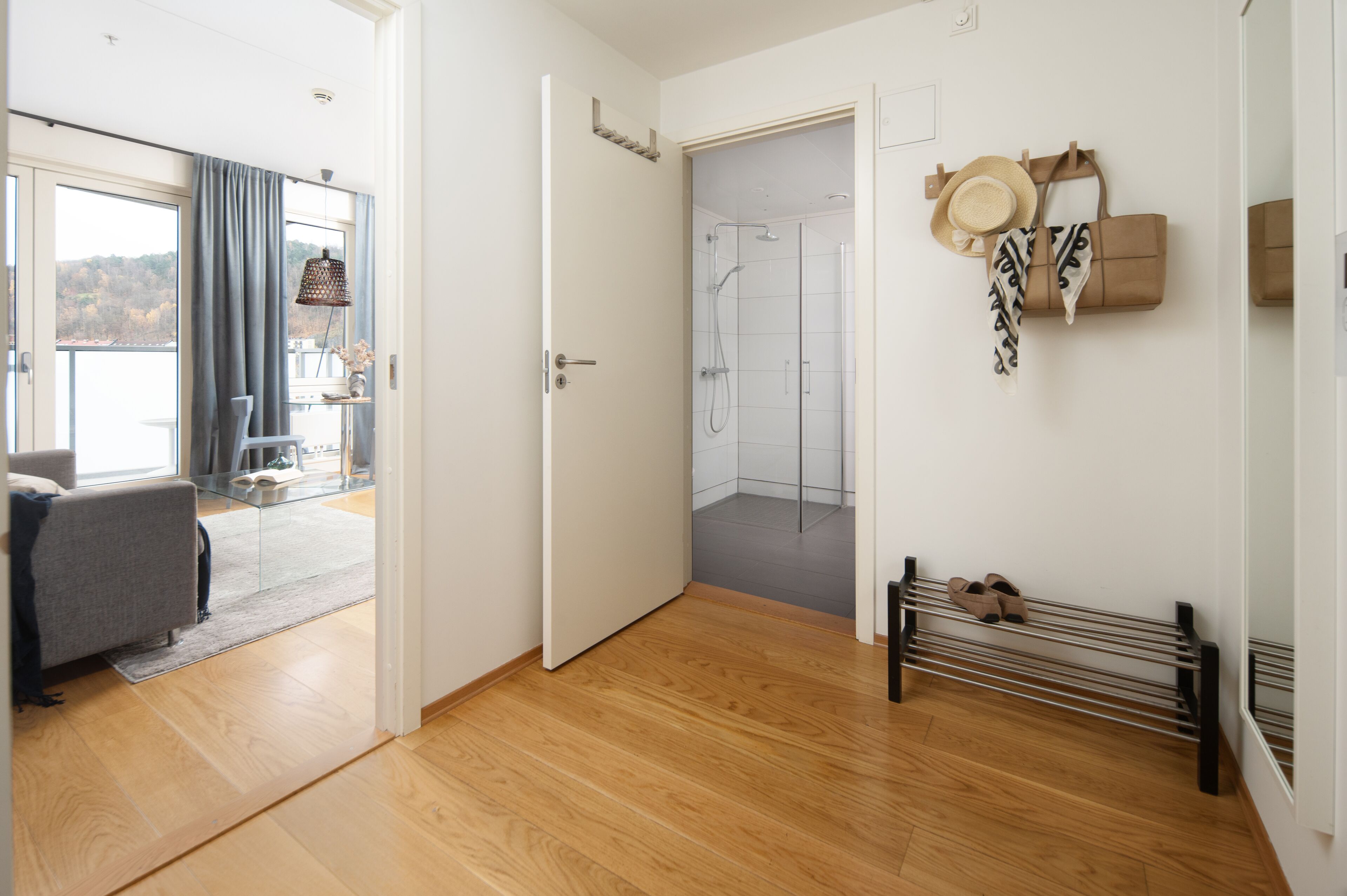 A modern hotel room with a wooden floor, a grey sofa, a glass shower enclosure, and a bench with shoes.