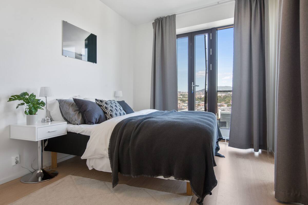 A modern bedroom with a bed, bedside tables, a mirror, and a view of the city through large windows.