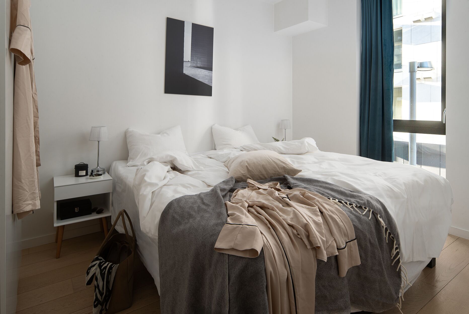 Cama bem arrumada com manta cinza e bege, mesa de cabeceira com abajur e janela com cortinas azuis.