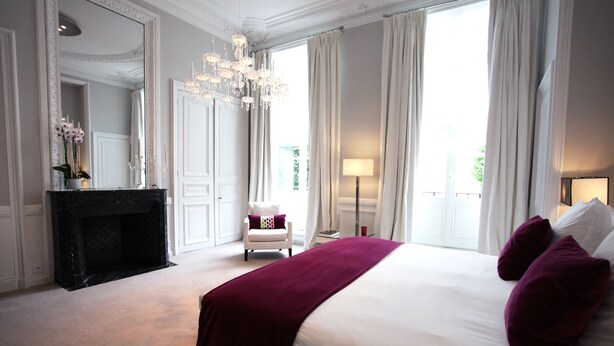 A spacious bedroom with a large bed, a fireplace, a chandelier, and a view of the outdoors. - Clarance Hotel Lille (Lille)