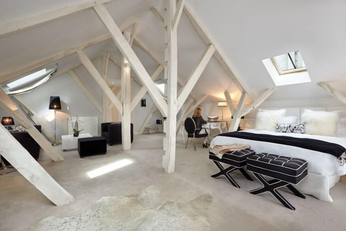 Un dormitorio en ático con techos altos y un techo inclinado, una cama con ropa de cama blanco y negro, un escritorio con una silla y una ventana pequeña.
