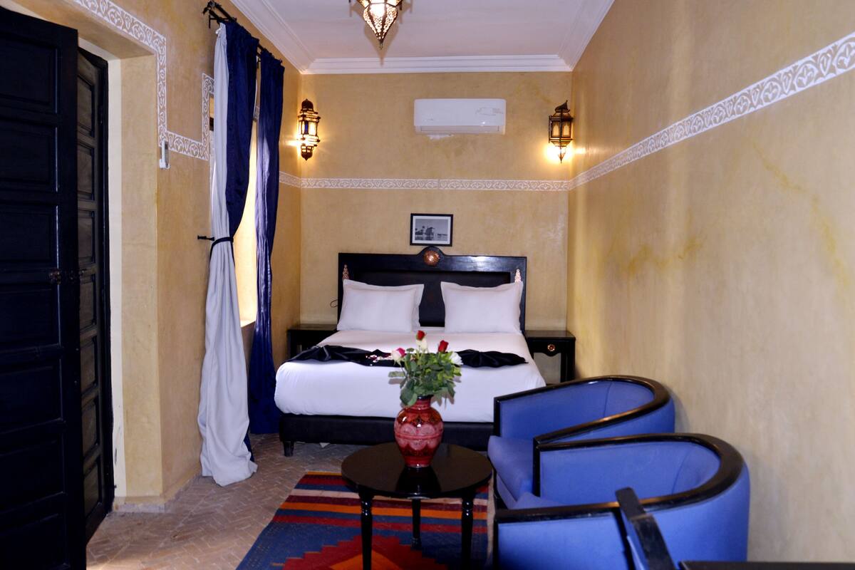 A hotel room with a bed, a small table with a vase of flowers, two blue chairs, and a black door.
